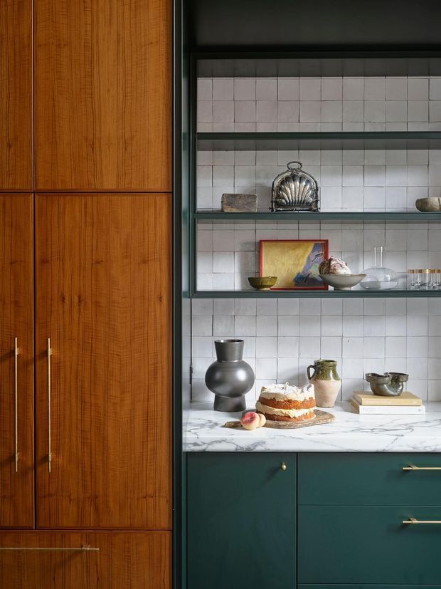 A richly styled kitchen storage wall featuring tall fluted mid-to-dark timber cabinetry with long brass handles on the left, deep green lower cabinetry on the right, open black-framed shelving, and a white marble-look benchtop. The shelving is backed with small square pale tiles and displays ceramics, glassware, artwork, and decorative objects. The composition has a refined, vintage-inspired character with strong material and colour contrast.