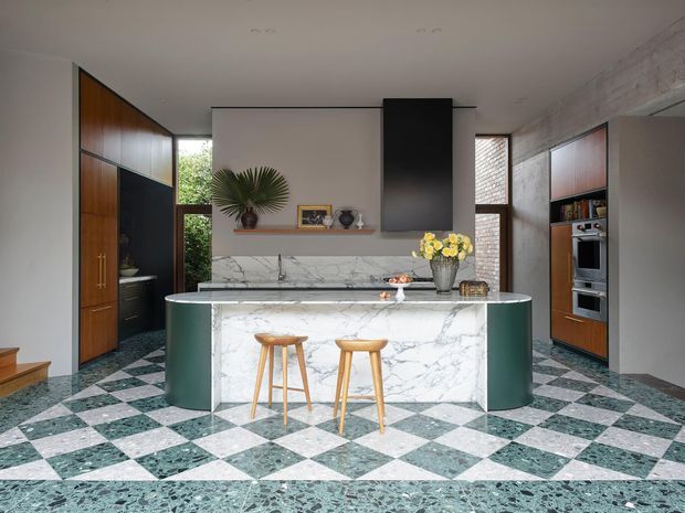 A sophisticated contemporary kitchen with a large marble-clad island, deep green curved cabinetry ends, dark timber and black built-in joinery, and a dramatic green-and-white checkerboard stone floor. The space includes integrated appliances, a marble splashback and benchtop, open shelving, a black rangehood, timber stools, and a mix of natural light and refined finishes.
