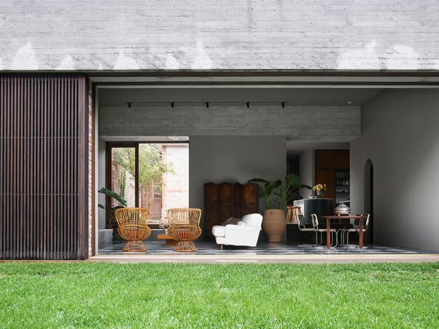 A modern concrete house facade opening onto a lawn, with a deep covered living area framed by dark timber or metal detailing. The interior features woven lounge chairs, a white armchair, timber cabinetry, indoor plants, and a small dining or bar table. Strong indoor-outdoor connection and restrained contemporary material palette.