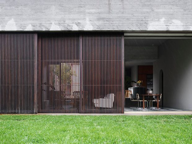 Front exterior view of a contemporary concrete residence with a deep recessed opening and a full-height dark timber slatted screen. The screen partially reveals an outdoor or semi-enclosed sitting area, while the open bay exposes a refined dining space with a timber table and chairs. A neatly maintained lawn occupies the foreground.