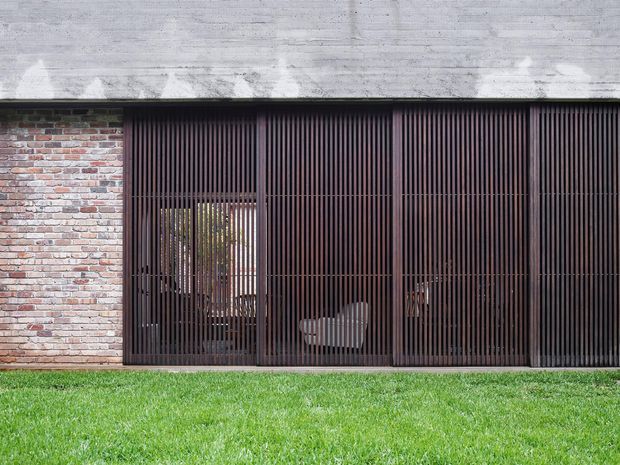 A minimalist contemporary building facade with a broad exposed concrete roof slab, weathered red-and-grey brick wall, and dark vertical metal screening across large openings. The screened facade partially reveals interior seating and planting behind it, with a neatly maintained lawn in front.