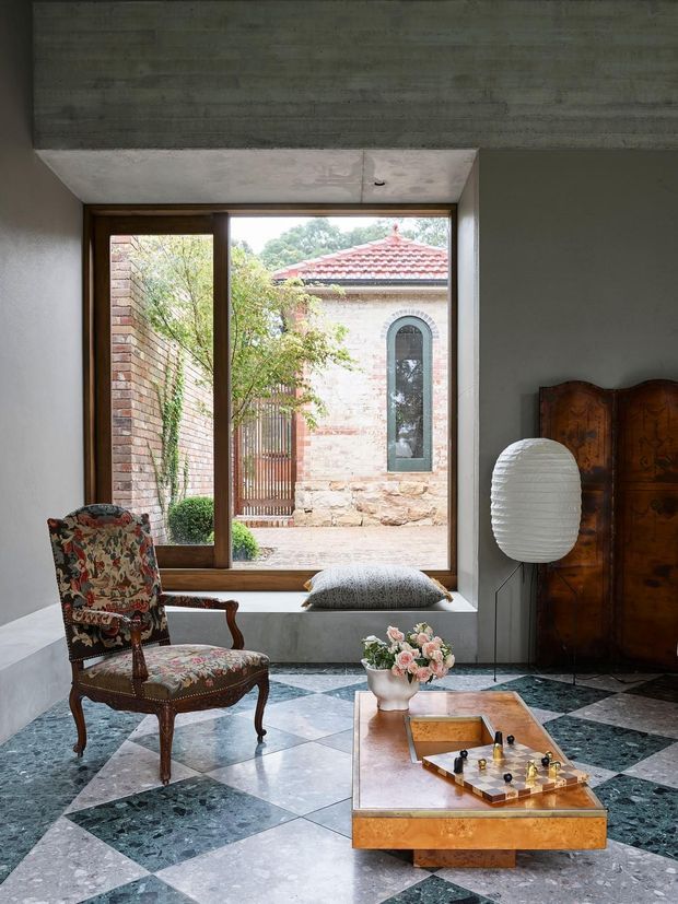 A refined living room with a large timber-framed window overlooking a brick courtyard, a patterned antique armchair, carved wooden armoire, sculptural floor lamp, and low timber coffee table. The room features a striking geometric marble or stone floor in dark green, grey, and white, with a concrete ceiling and neutral walls.