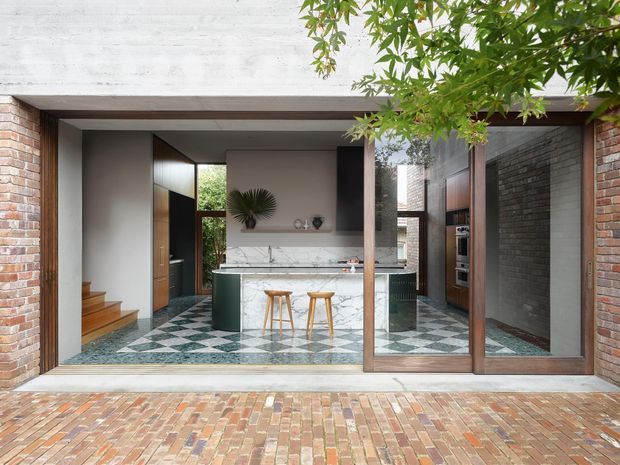 View through a large glazed opening into a refined contemporary kitchen. The space features exposed brick walls, a dramatic geometric patterned tile floor, dark green cabinetry, a white marble kitchen island with integrated seating, timber stools, dark timber-framed glazing and an adjacent stair. A paved brick terrace and leafy planting are visible outside.