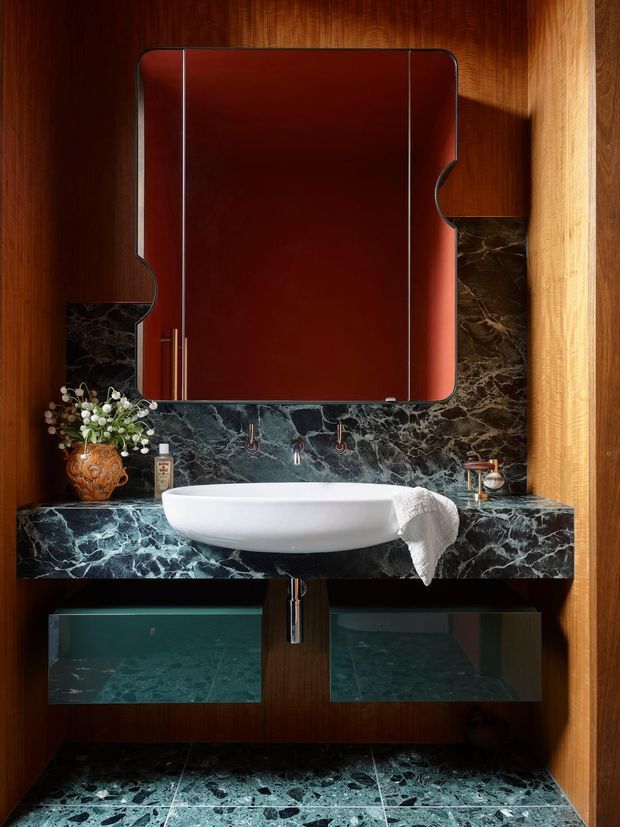 A dramatic, richly detailed bathroom vanity featuring a white vessel basin on a dark green-black veined stone benchtop, matching stone backsplash and floor tiles, dark timber wall panelling, and a large red-brown framed mirror. Floating dark timber cabinetry and exposed plumbing sit beneath the vanity, with warm directional lighting and decorative accessories.