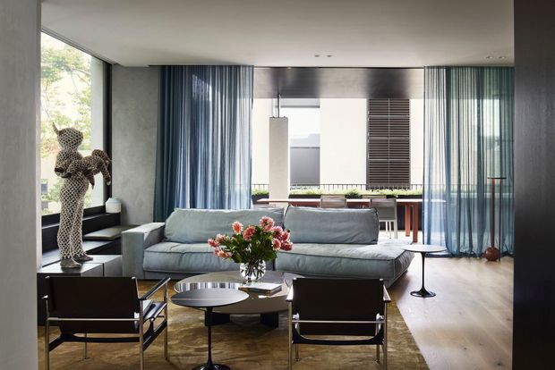 A refined contemporary living room with a large low-profile grey sectional sofa, black-framed lounge chairs, round nesting coffee tables, and a floral arrangement over a textured rug. Full-height sheer blue-grey curtains frame extensive glazing, while a sculptural spotted artwork stands on a side console. The room opens visually toward a dining or outdoor area beyond, with warm timber flooring and a restrained neutral palette.