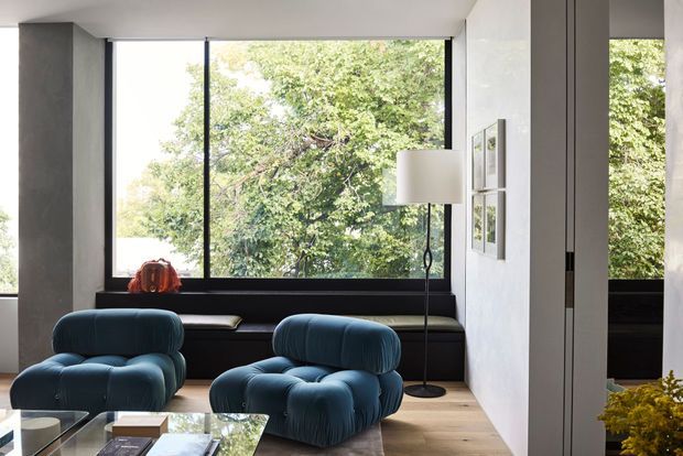 Contemporary living room with two deep blue sculptural lounge chairs, a low glass coffee table, pale timber flooring, and a dark built-in window seat beneath expansive black-framed glazing. A floor lamp and framed artwork sit against white walls, with leafy greenery visible outside.