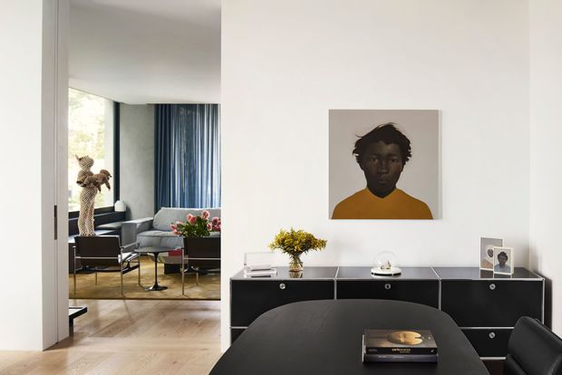 Contemporary dining area with a black dining table and chairs, a long black sideboard, and a large portrait artwork on a white wall. Through the wide opening is a connected sitting area with blue curtains, upholstered seating, a rug, and a tall indoor plant. The space has pale timber flooring, clean white walls, and restrained modern furnishings.