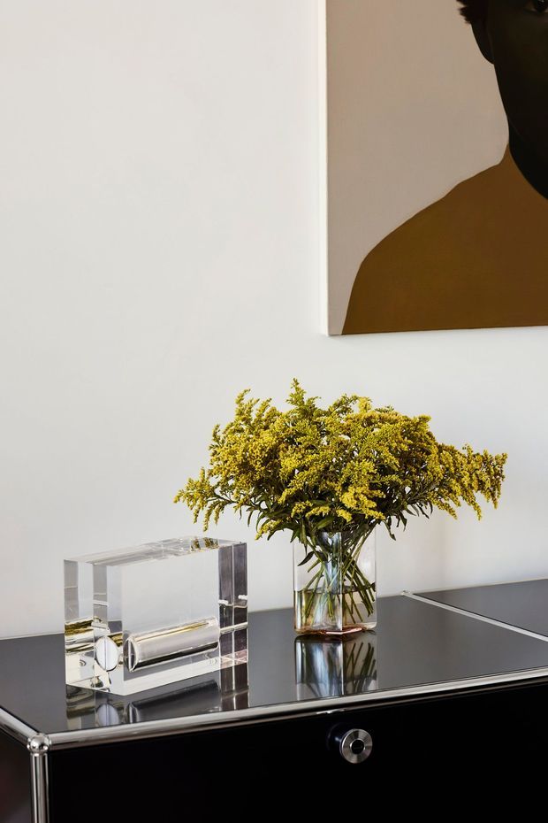 A close-up interior vignette featuring a glossy black console or sideboard with chrome detailing, a clear acrylic decorative object, and a glass vase of yellow flowers. A large abstract artwork with a dark brown and black figure hangs on the white wall above. The composition is refined and high-contrast, with reflective surfaces and restrained contemporary styling.