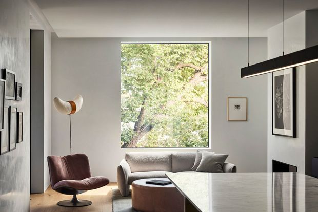 A refined contemporary living room with a large picture window overlooking trees, sculptural lounge seating, a curved coffee table, framed artwork, and a long linear pendant light. A substantial stone island or counter occupies the foreground, creating a polished, gallery-like interior with a restrained neutral palette.