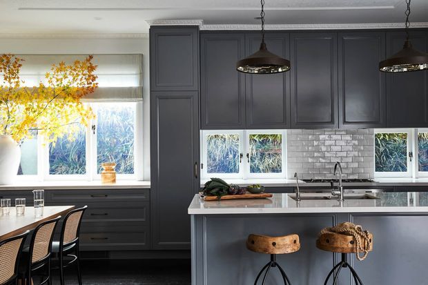 A refined contemporary kitchen with dark charcoal-grey cabinetry, a large central island with a pale stone benchtop, white subway-tile splashback, and black pendant lights. The space includes a compact dining area with woven-back bar stools and abundant natural light through multiple windows.