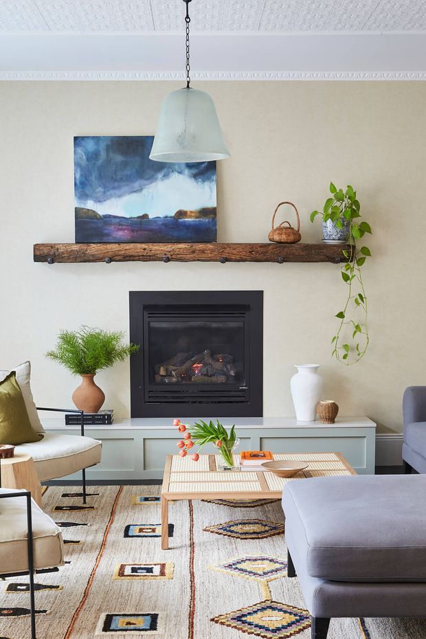 A warmly styled living room featuring a black built-in fireplace set into pale cabinetry, a rustic timber mantel shelf, upholstered seating, a low timber coffee table, patterned area rug, pendant light, plants, and artwork. The room combines contemporary furnishings with rustic and eclectic decorative elements.