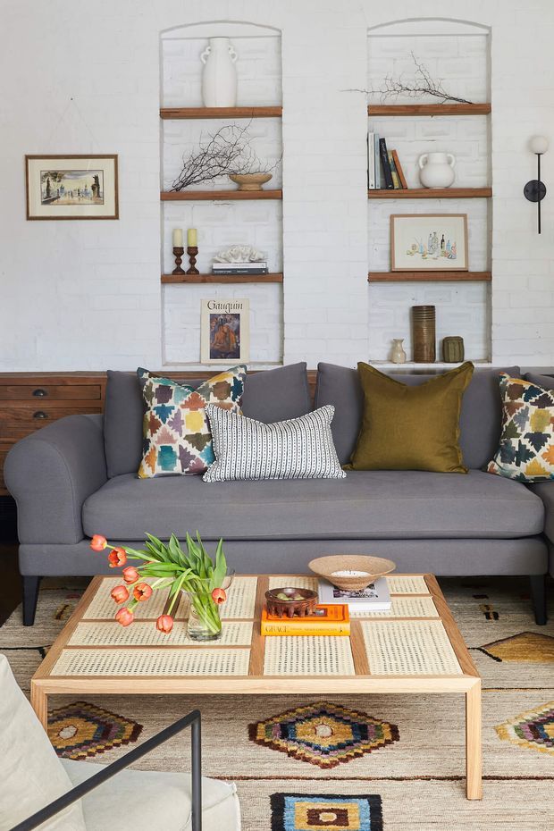 A styled living room featuring a large charcoal-grey sofa with colourful patterned and olive cushions, set against a white-painted brick wall with recessed timber display shelves. The shelves hold ceramics, books, artwork and natural branch arrangements. A light timber coffee table with woven inset panels occupies the foreground, placed over a neutral rug with multicoloured geometric motifs. The room has a curated, eclectic and warm atmosphere with strong textural and handcrafted elements.