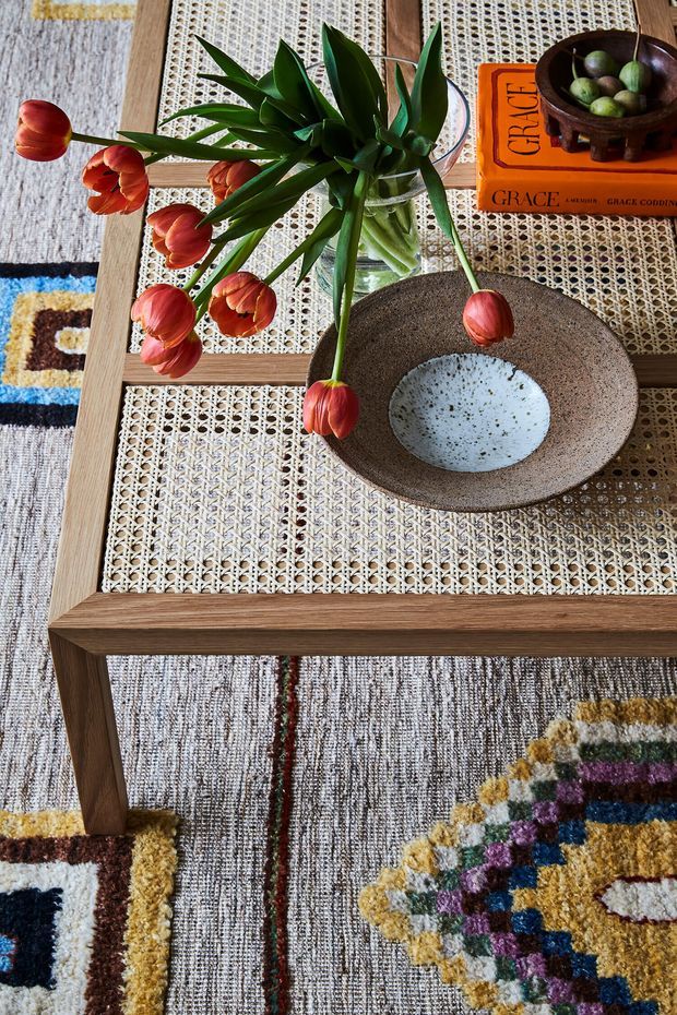 A close-up, overhead view of a styled living-room coffee table with a timber frame and woven cane inset top. A clear glass vase holds red tulips, beside a rustic ceramic bowl, an orange book, and a small wooden bowl of green fruit. The table sits on a richly patterned, multicoloured textured rug.