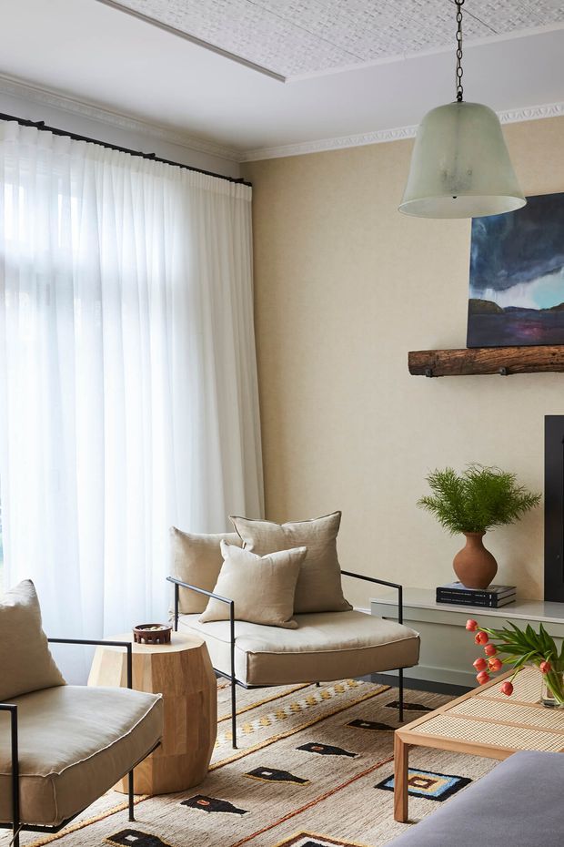 Bright, refined living room with tall sheer curtains, cream upholstered lounge chairs, a patterned geometric rug, a timber coffee table, and a dark metal-framed armchair. The room features a textured white ceiling, decorative cornices, a large pendant light, a rustic timber mantel shelf, artwork, and indoor plants.