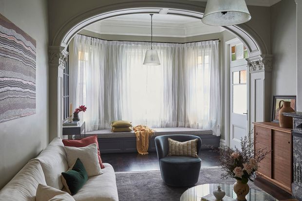A refined living room with a deep bay window framed by sheer curtains, an upholstered sofa, sculptural armchair, circular coffee table, built-in window seat, timber cabinetry and an ornate arched opening with decorative classical detailing. The room is softly lit and styled in a muted, elegant palette.