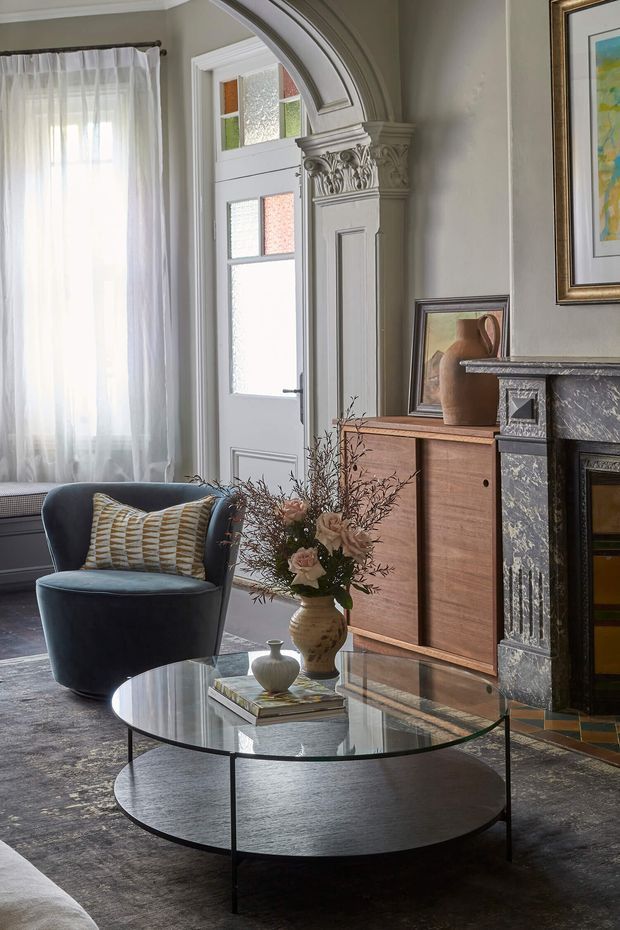 A refined living room with a tall sheer-curtained window, ornate white entry door and architrave, decorative carved column detail, dark marble fireplace, timber sideboard, sculptural armchair, and round glass coffee table. The room combines heritage architectural detailing with contemporary furnishings and muted neutral tones.