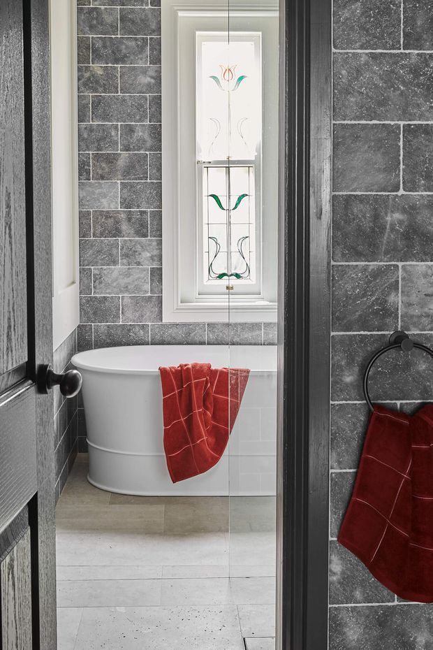 A compact bathroom viewed through a partially open dark timber door, featuring charcoal-grey brick-pattern wall tiles, pale stone floor tiles, a white freestanding bathtub beneath a narrow decorative stained-glass window, and a vivid red towel. A mirror on the right reflects the towel rail and tiled wall.