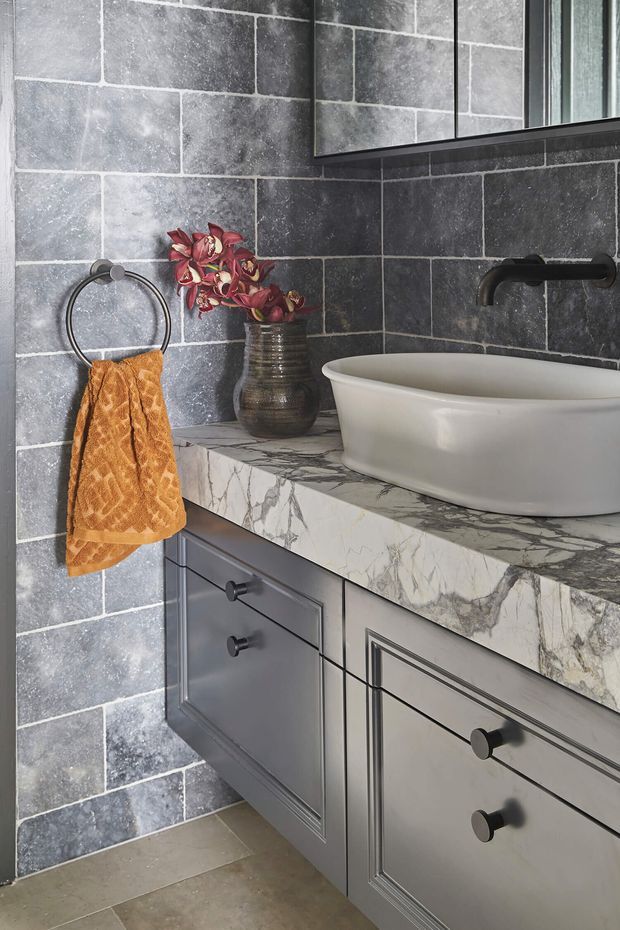Close-up view of a refined bathroom vanity with a white vessel basin, dark wall-mounted tapware, grey-blue framed cabinetry, and a veined white natural-stone benchtop. Large charcoal-grey rectangular wall tiles with light grout create a textured backdrop, while a bronze towel and decorative vase add warmth. A mirror is visible above the vanity.