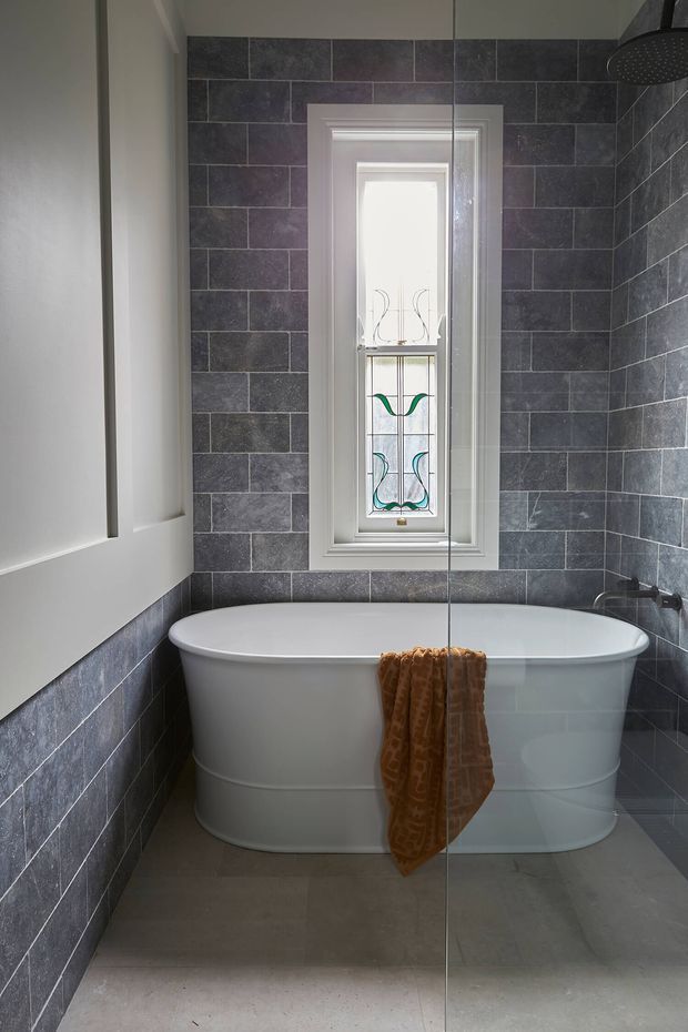 A compact, refined bathroom with a freestanding white bathtub positioned beneath a narrow stained-glass window. Dark charcoal-grey rectangular wall tiles wrap the room, contrasting with white painted trim and a white built-in cabinet. A frameless glass shower screen and wall-mounted shower fixture are visible at right, with large grey floor tiles underfoot.