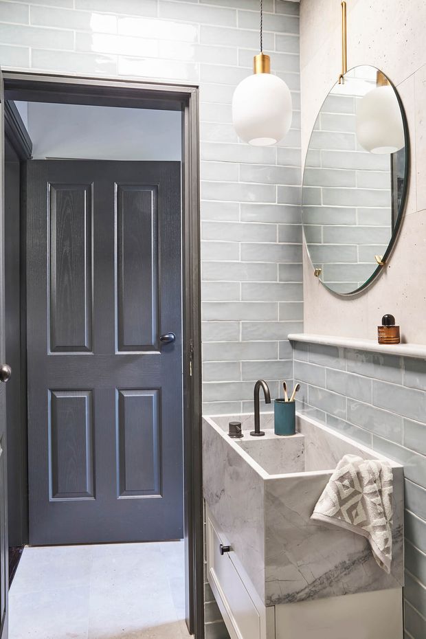 A compact, refined bathroom featuring pale grey-green glossy subway wall tiles, a white marble-look vanity with integrated rectangular basin, dark charcoal panelled door, oval wall mirror, and a white globe pendant with brass fitting. The palette is light and cool with contrasting dark joinery and warm metallic accents.