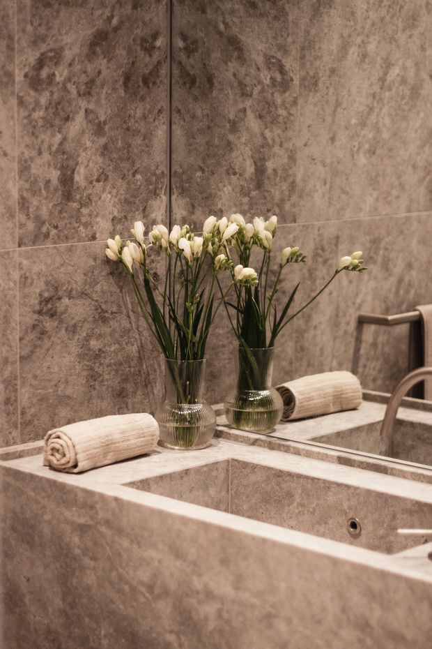 Close-up view of a refined bathroom vanity and bathtub area, featuring continuous warm taupe stone-look wall and bath surfaces, a large mirror, rolled cream towels, and clear glass vases with white flowers. A chrome towel rail and partial basin fittings are visible at the right.