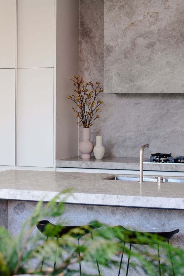 A refined contemporary kitchen featuring pale grey stone surfaces, a large island with an integrated sink and minimalist chrome tap, full-height white cabinetry, and a matching stone splashback with a raised feature panel. A gas cooktop is built into the rear benchtop, while sculptural ceramic vases and foliage add subtle styling. Dark stools are partially visible beneath the island.