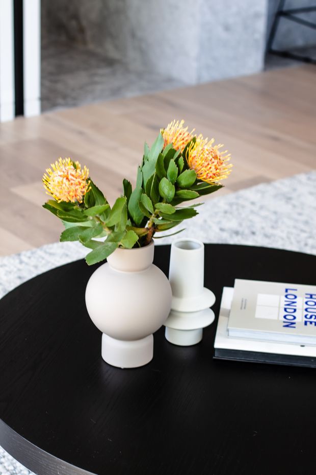 Close-up interior styling vignette featuring a round black coffee table with a matte white sculptural vase holding orange-yellow flowers, matching white ceramic vessels, and stacked design books. The setting includes pale timber flooring, a light textured rug, a grey concrete-like wall, and partial black metal furniture in the background.