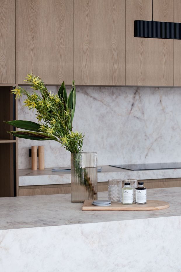Close-up view of a refined contemporary kitchen featuring warm light-wood cabinetry, a pale veined stone splashback and island benchtop, dark integrated appliances or joinery accents, and a sculptural black linear pendant light. A ribbed glass vase with greenery, glassware, and small jars sit on a timber tray atop the island.