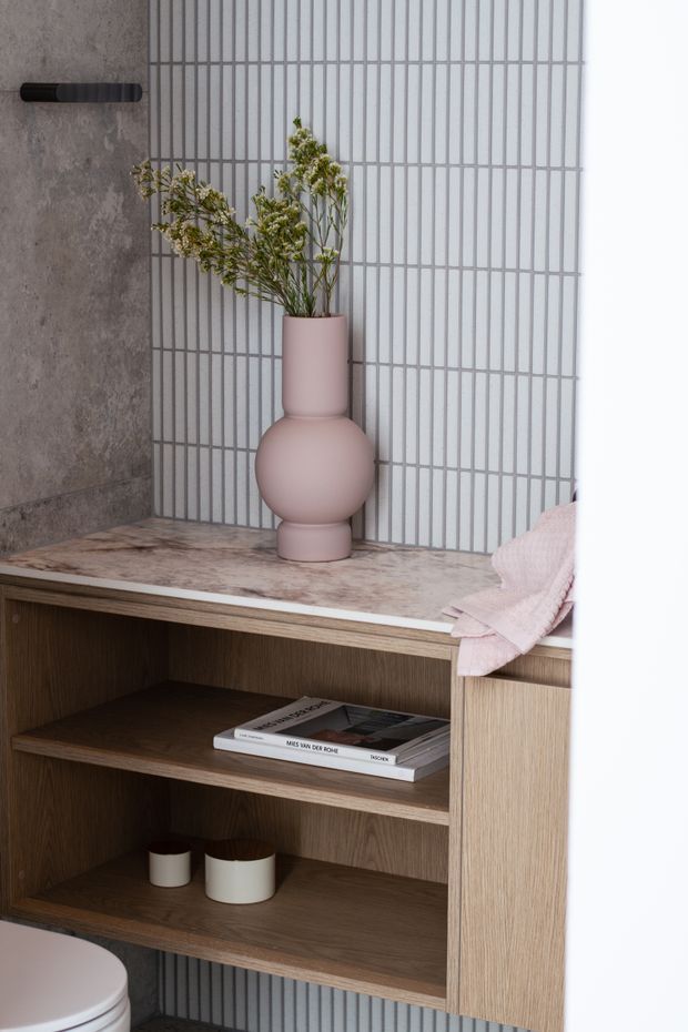 Close-up of a refined bathroom vanity with open dark timber shelving, a pale stone or marble benchtop, vertical grid-pattern tiled splashback, and a soft pink sculptural vase with greenery. A partial toilet and towel are visible, creating a warm, contemporary bathroom vignette.