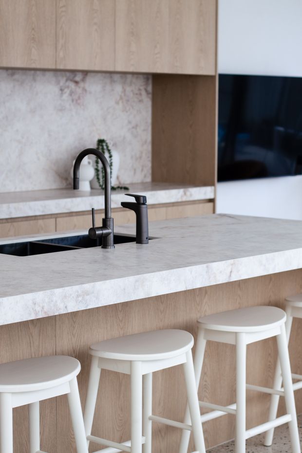 Contemporary kitchen interior featuring a large light stone island with waterfall-style edging, integrated black sink and dark tapware, light timber cabinetry, a pale stone or marble-look splashback, and white bar stools. A television is mounted on the wall beyond the island.