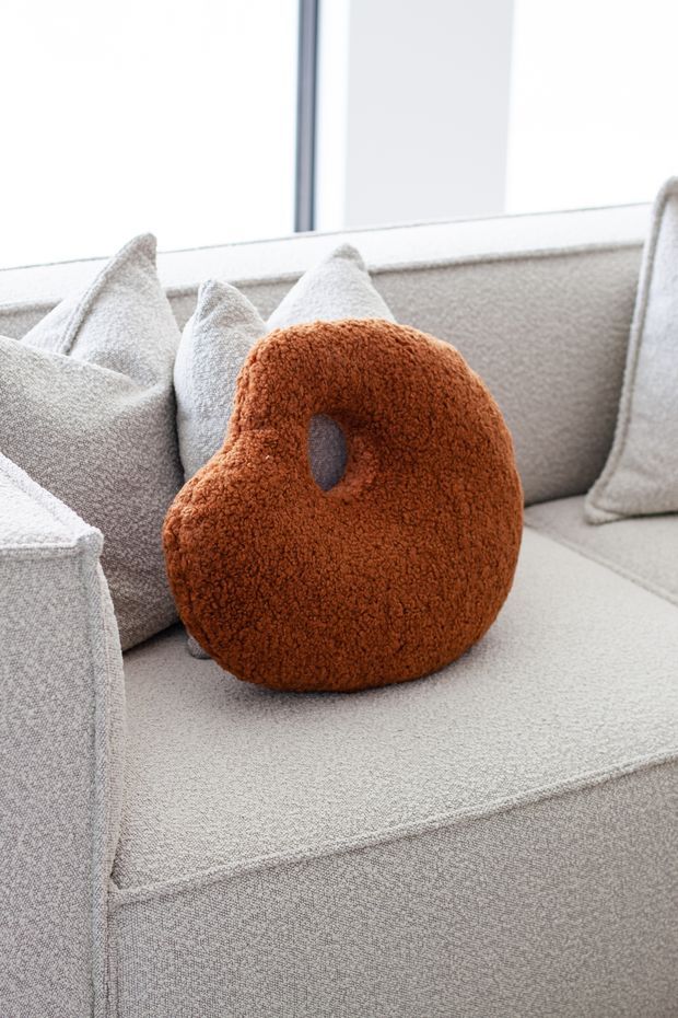 Close-up of a contemporary upholstered sofa in a bright, neutral-toned interior. The sofa is layered with light grey textured cushions and features a prominent rust-orange boucle accent cushion with an oval cut-out detail. Large glazing in the background provides soft natural light.