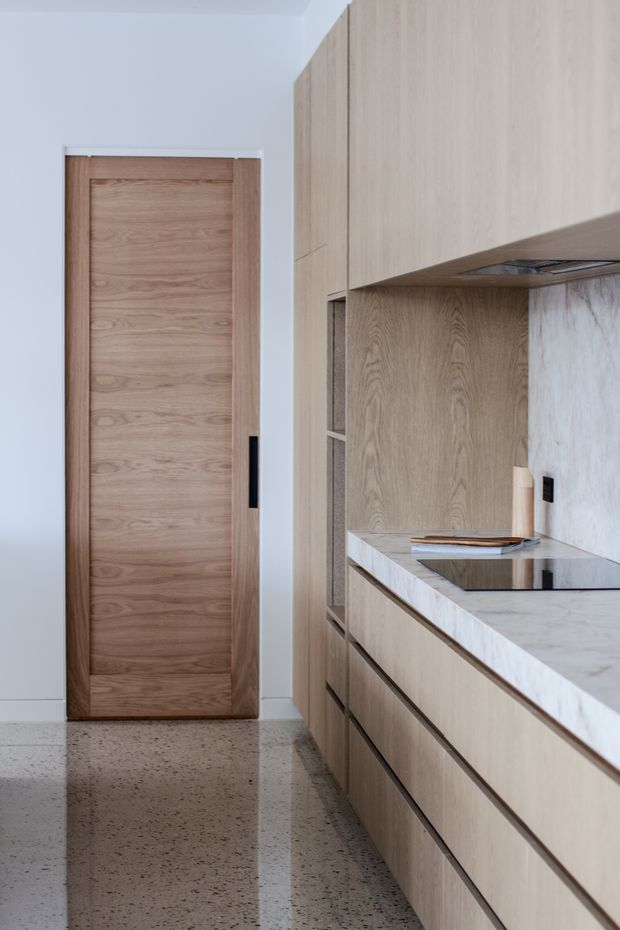 A narrow, refined contemporary kitchen viewed along a run of light timber cabinetry. The space features full-height wood-grain cupboards, integrated lower drawers, a pale stone benchtop with an induction cooktop, a textured light stone or plaster splashback, and a matching timber internal door. The floor is finished in grey terrazzo or polished concrete with dark aggregate. Minimal black hardware provides subtle contrast.