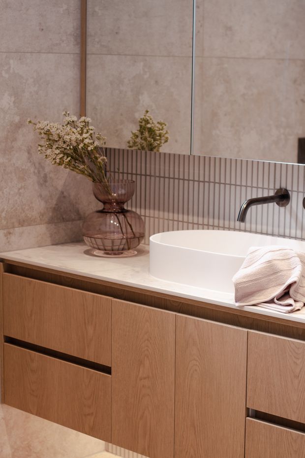 A refined contemporary bathroom vanity featuring warm wood-grain cabinetry, a pale stone benchtop, a white countertop basin, brushed dark metal wall-mounted tapware, large beige stone-look wall tiles, and a narrow vertical mosaic tile splashback. A frameless mirror and decorative glass vase complete the warm, luxurious composition.