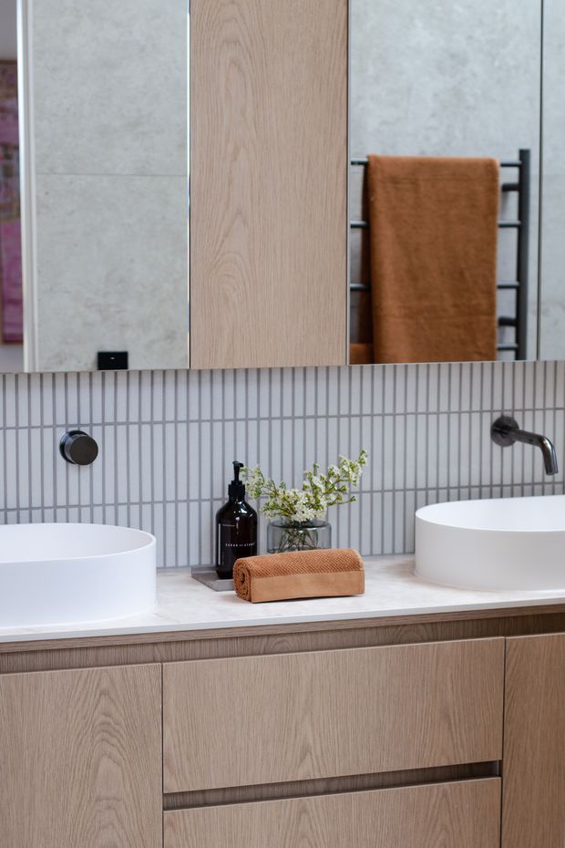 A contemporary bathroom vanity with two white vessel basins set on a pale stone benchtop, warm light-oak cabinetry, and a vertically lined grey tile splashback. Black wall-mounted tapware and a black heated towel rail contrast with the neutral finishes, while a rust-brown towel and small plant add warmth. A large mirror and timber storage cabinet are visible above the vanity.