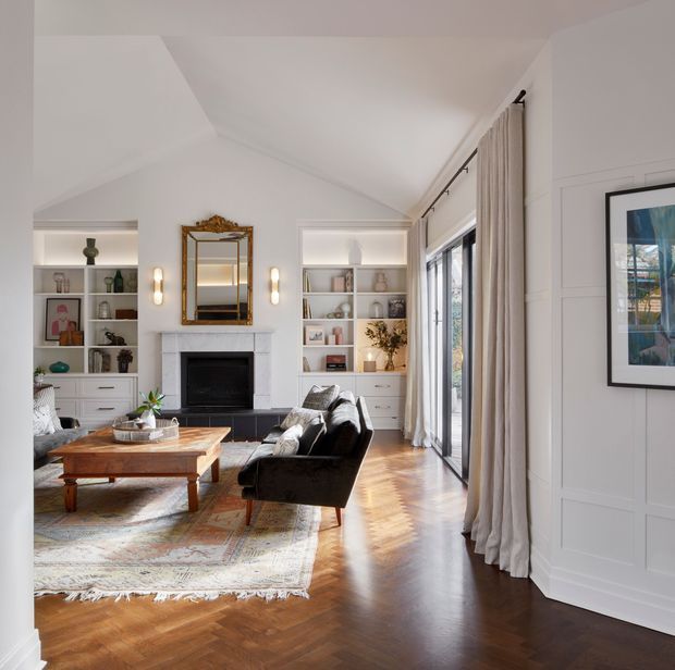 A refined living room with built-in white shelving flanking a traditional fireplace, an ornate framed mirror, black sofas, a substantial timber coffee table, and a patterned area rug. Herringbone timber flooring, white wall panelling, tall windows with full-length curtains, and layered display styling create a bright yet elegant interior.