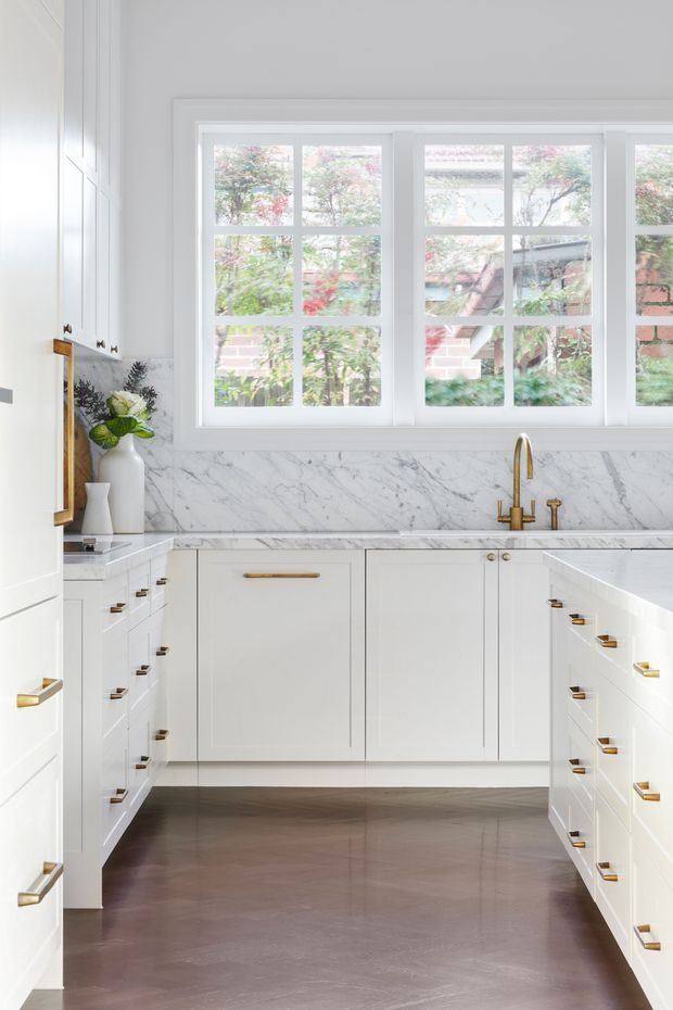 Bright, refined white galley-style kitchen with extensive shaker-profile cabinetry, brass pulls and faucet, white marble benchtops and full-height marble splashback. Large multi-pane windows provide abundant natural light and overlook greenery and a brick exterior. Dark timber flooring contrasts with the predominantly white fit-out.