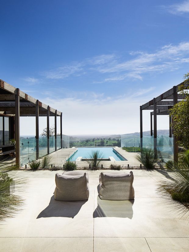 A bright outdoor terrace overlooking a rolling green landscape, with a rectangular infinity-edge pool, glass balustrades, timber pergola structures, concrete paving, sculptural grasses and two neutral lounge chairs. The scene has a contemporary resort-like feel and expansive rural views.