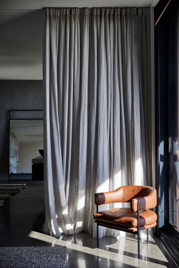 A moody, refined interior corner with full-height sheer grey curtains, dark framed floor-to-ceiling glazing, polished dark flooring, and a sculptural cognac leather lounge chair. Strong sunlight creates dramatic shadows across the floor, while a large mirror reflects the adjoining dark-toned space.