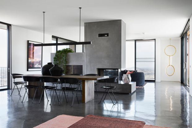 Open-plan contemporary living and dining area with a long timber dining table, black wire-frame chairs, polished concrete floors, extensive floor-to-ceiling glazing, and a sculptural concrete fireplace or media feature. The room is bright and minimalist, with ocean or coastal scenery visible beyond the windows and a small seating area near the fireplace.