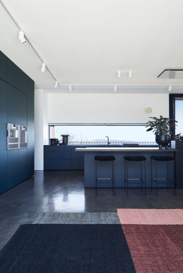 A contemporary kitchen with dark blue built-in cabinetry, a large island with a pale stone benchtop and three stools, polished dark flooring, and a wide window overlooking a distant landscape. White walls and ceiling-mounted track lighting create a bright, clean interior, with colourful area rugs and a potted plant adding contrast.