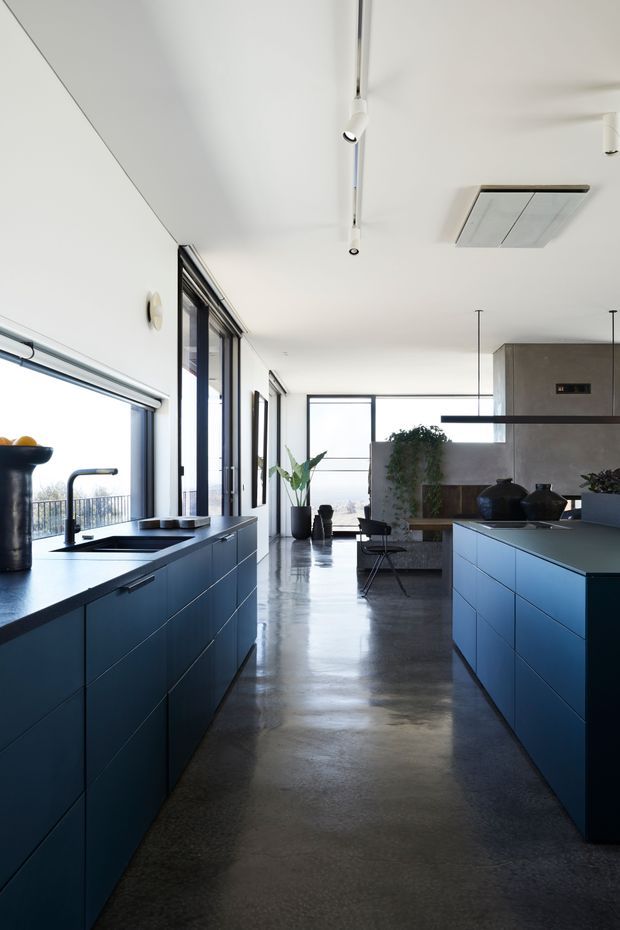 A bright, open-plan contemporary kitchen with two parallel banks of deep blue cabinetry and dark benchtops. The kitchen includes a sink beneath expansive glazing, integrated appliances, a central walkway, polished concrete flooring, black-framed windows and doors, and a dining or living area beyond. Track lighting, a large ceiling-mounted air-conditioning unit, indoor plants and minimalist furnishings reinforce the clean modern character.