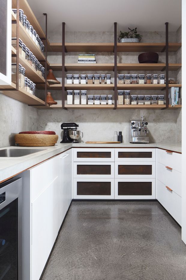 A compact, highly organised kitchen or pantry with white lower cabinetry, dark timber drawer fronts and leather pulls, open timber shelving on dark metal brackets, and a textured grey stone or concrete backsplash. The shelves are filled with labelled jars and kitchen supplies. A sink, stand mixer, espresso machine, and under-counter appliance are visible. The space has a warm, functional contemporary design with industrial-inspired shelving.