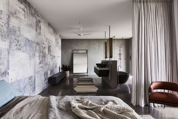 A sophisticated open-plan bedroom with a dark polished floor, textured abstract feature wall, freestanding black bathtub, floating vanity, and full-height sheer curtains. A partially visible bed occupies the foreground, while a sculptural brown leather chair and ceiling fan add warmth and functionality. The palette is predominantly grey, taupe, black, and brown with a refined contemporary feel.