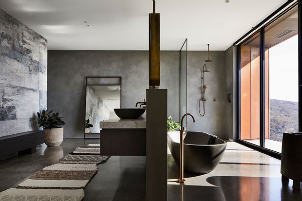 A dramatic, contemporary bathroom with a freestanding dark bathtub, sculptural basin, open shower and floating vanity. The room features textured grey concrete-like walls, dark polished flooring, frameless glass shower panels, black and bronze fixtures, and expansive floor-to-ceiling glazing overlooking a mountainous landscape. A long linear pendant adds a strong vertical architectural feature.