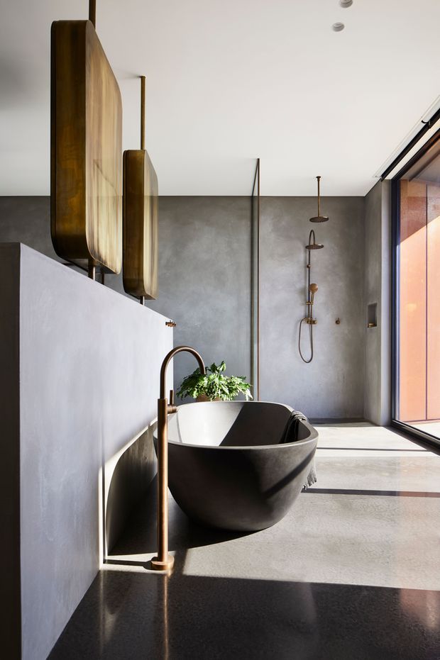 A contemporary bathroom featuring a freestanding dark stone bathtub, wall-mounted shower fittings, a tall bronze-toned bath tap, polished dark stone vanity surface, grey plaster walls, and large full-height glazing. Sculptural bronze pendant lights and a potted plant add warmth to the minimalist space.