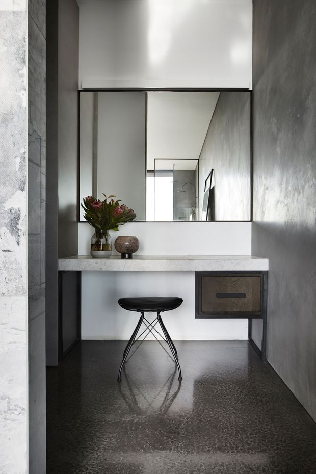 A compact, refined vanity or dressing area with a floating stone-look benchtop, large black-framed mirror, dark textured walls, polished dark flooring, and a small built-in timber drawer. A black wire-frame stool and floral arrangement add contrast. The mirror reflects a nearby bathroom or shower area.