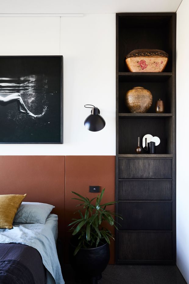 A refined bedroom corner with a partially visible bed, rust-toned upholstered wall panel and headboard, white walls, and a tall dark timber built-in shelving and drawer unit. The shelving displays decorative ceramics and objects, complemented by a black wall sconce, monochrome artwork, blue-grey bedding, and a potted plant. The palette is warm, dark, and contemporary with strong contrast.