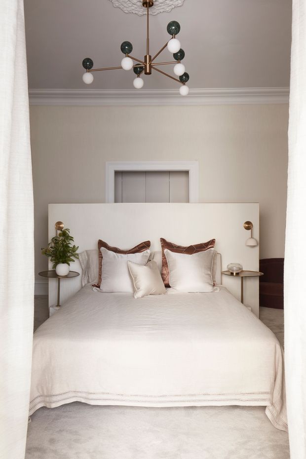 A refined bedroom viewed through sheer curtains, featuring a neatly made upholstered bed with layered cream and white bedding, a low pale headboard, slim bedside tables, and integrated bedside wall lights. A sculptural brass-and-glass pendant hangs from an ornate ceiling medallion. The room has softly painted walls, detailed cornicing, a concealed doorway behind the bed, and a muted, luxurious neutral palette.