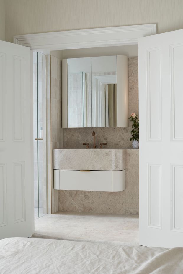 A refined bathroom viewed directly from a bedroom through open white paneled double doors. The bathroom features a floating white vanity with a thick beige natural-stone benchtop, matching stone wall and floor surfaces, bronze-toned tapware, mirrored wall cabinets, and a glass shower enclosure. Traditional wall mouldings and restrained contemporary fixtures create a polished, elegant appearance.