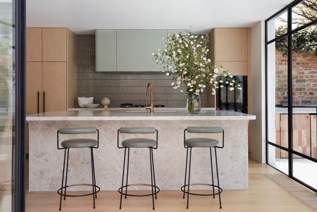 A refined contemporary kitchen centred on a large pale stone island with seating for three. Full-height light timber cabinetry frames muted green overhead cabinetry and a dark tiled splashback, with a copper-toned tap and integrated appliances. Black-framed glazing opens to a brick exterior, while light timber flooring and soft neutral finishes create a warm, elegant interior.